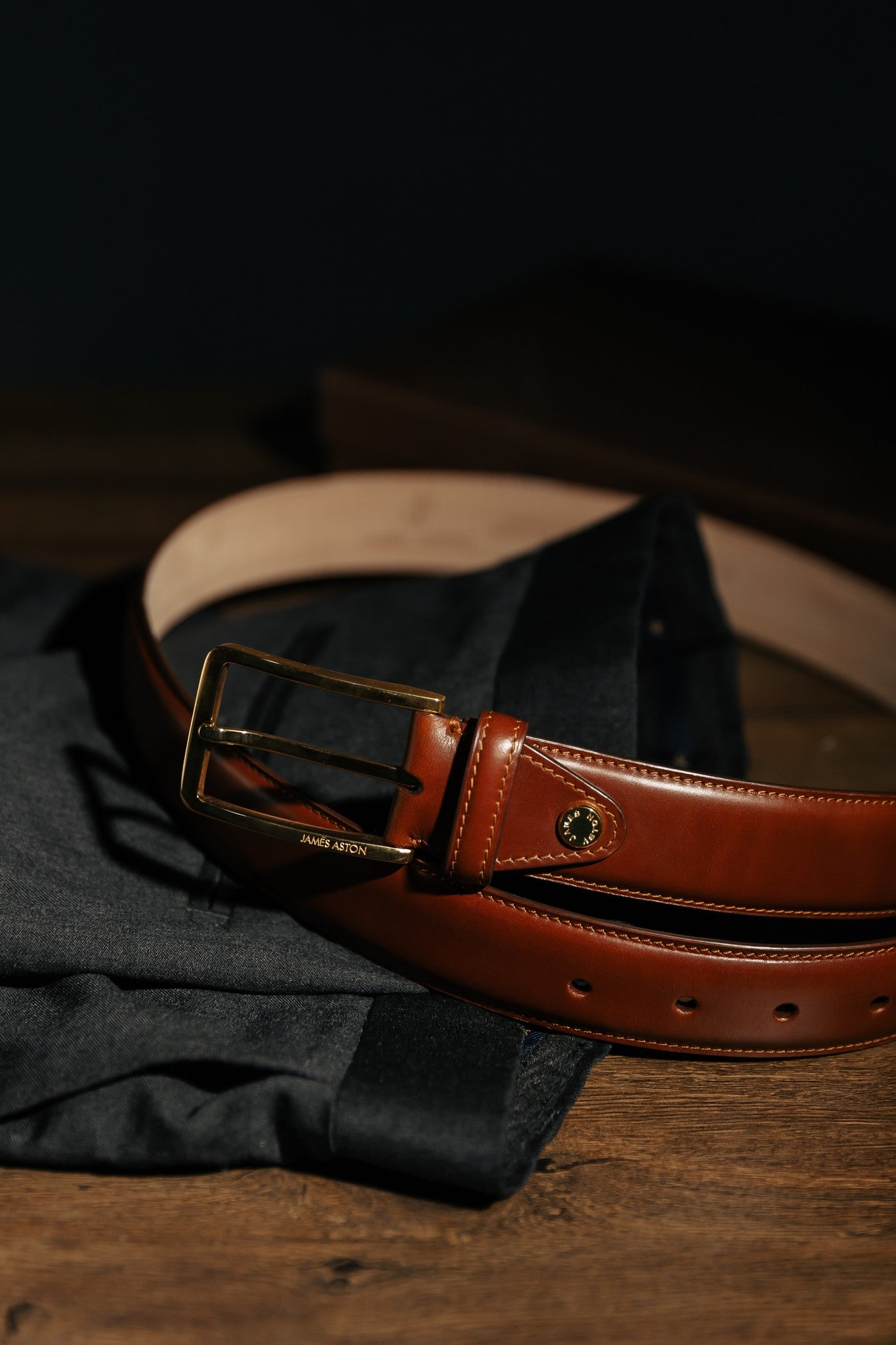 The Lorenzo cognac Italian Bridle leather belt at rest on folded charcoal cloth, by James Aston