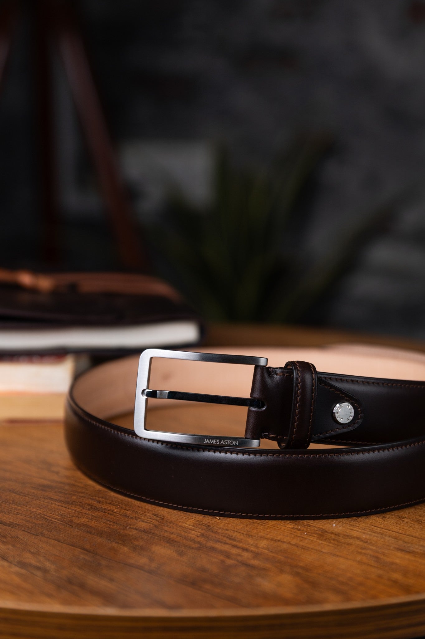 The Lorenzo dark brown Italian Bridle leather belt at rest on a wooden surface, by James Aston