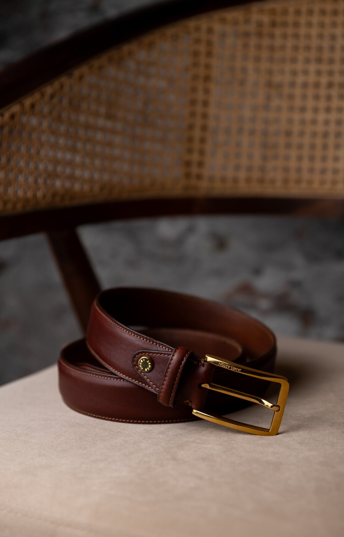The Raffaello oak brown milled Vachetta leather belt resting against a cane chair in warm light