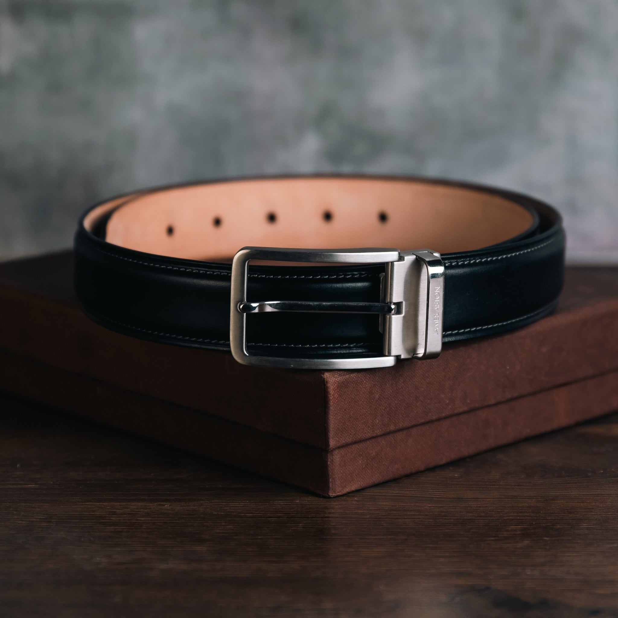 The Cesare black Italian Vegetable Tanned Bridle leather belt coiled on dark wood, nickel buckle and natural honey nubuck lining, James Aston